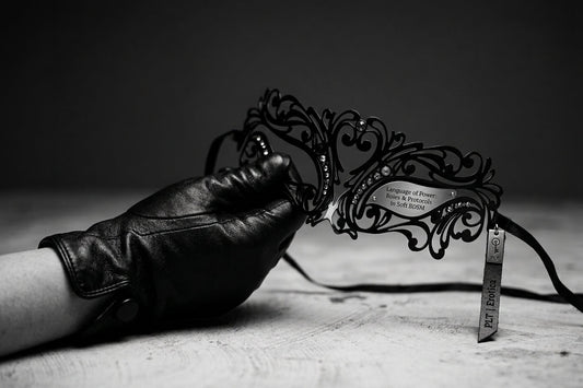 A close-up shot of a gloved hand holding an ornate black lace masquerade mask, symbolizing soft BDSM roles and protocols.