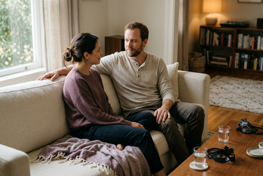 Couple sharing a quiet moment on a cream sofa, with a soft blanket beside them and masquerade masks and water glasses on the coffee table.