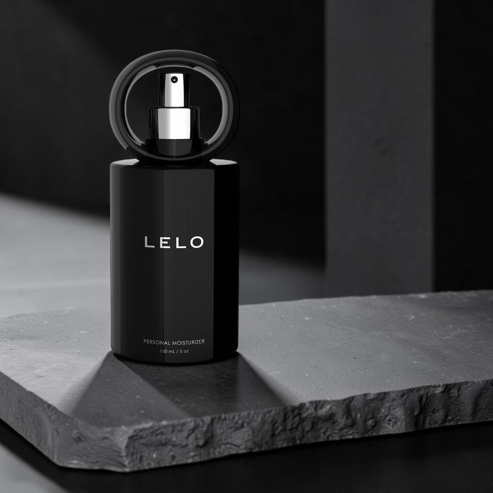LELO Personal Moisturizer perfume bottle in matte black with a circular black cap and silver spray nozzle, displayed on a dark textured stone slab against a dark gray background.
