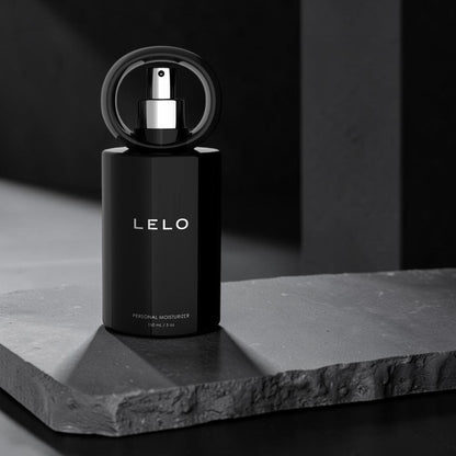 LELO Personal Moisturizer perfume bottle in matte black with a circular black cap and silver spray nozzle, displayed on a dark textured stone slab against a dark gray background.