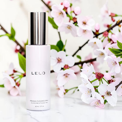 A bottle of LELO Premium Cleaning Spray standing upright on a light marble-like surface, surrounded by blooming pink and white cherry blossoms with green leaves.