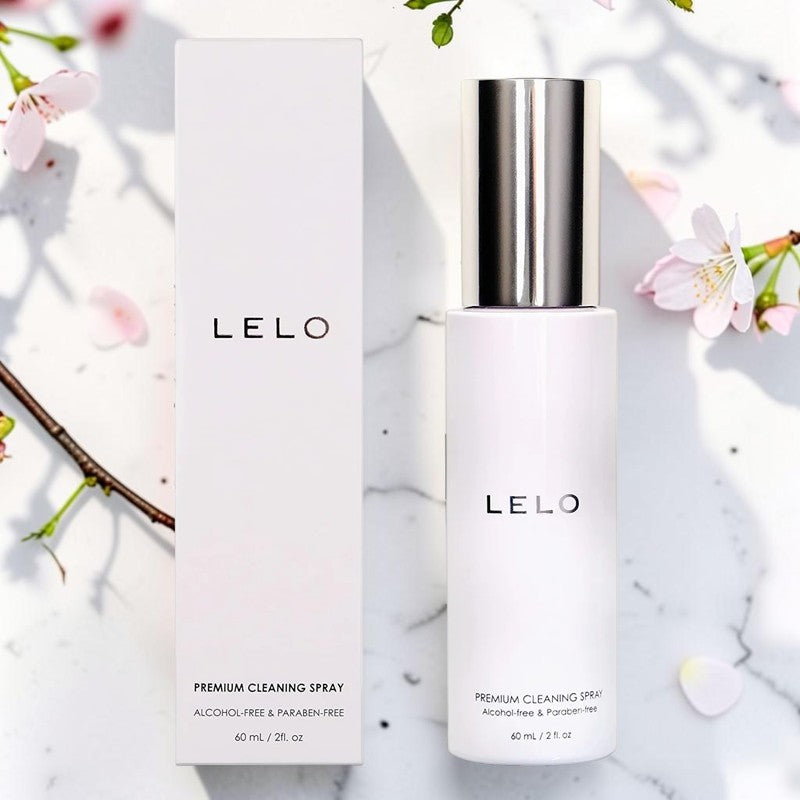 LELO Premium Cleaning Spray bottle with a metallic silver cap next to its white packaging box, styled on a white marble surface with cherry blossom petals.