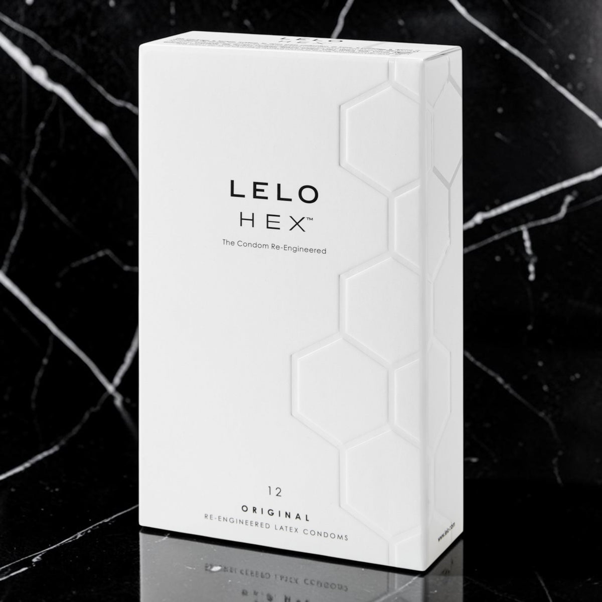 A white rectangular box of LELO HEX condoms stands upright on a glossy black marble surface with a dark marble background. The box has a subtle embossed hexagon pattern and black text reading 'LELO HEX' and '12 ORIGINAL RE ENGINEERED LATE CONDOMS'.
