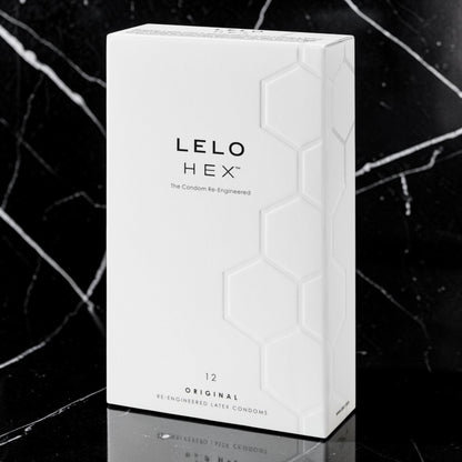 A white rectangular box of LELO HEX condoms stands upright on a glossy black marble surface with a dark marble background. The box has a subtle embossed hexagon pattern and black text reading 'LELO HEX' and '12 ORIGINAL RE ENGINEERED LATE CONDOMS'.