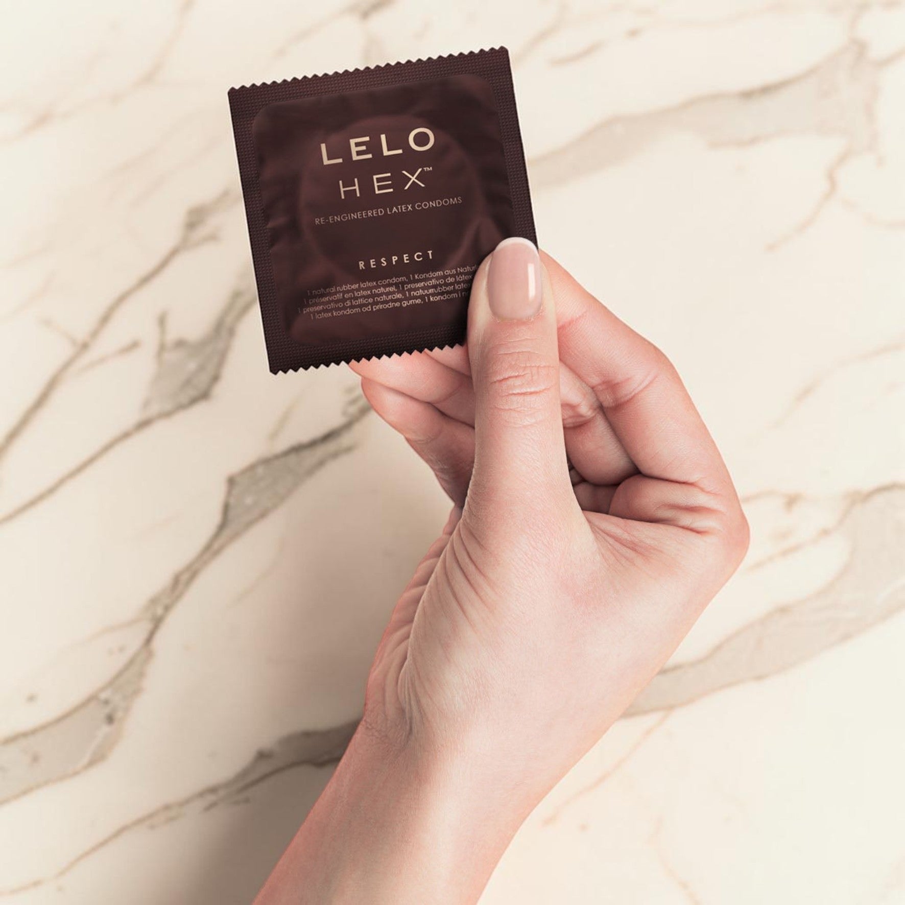 A hand holding a small dark brown LELO HEX lubricant packet against a light beige marble background.